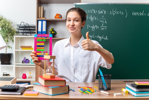 pleased young female math teacher sitting at desk with school supplies holding abacus looking at front showing thumb up in classroom