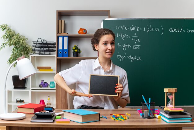 교실 앞을 바라보는 미니 칠판을 들고 가리키는 학용품을 들고 책상에 앉아 있는 젊은 여성 수학 교사를 기쁘게 생각합니다