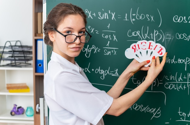 교실 앞을 보고 있는 러시아 알파벳 팬을 들고 칠판 앞에 프로필 보기에 서 있는 안경을 쓴 젊은 여성 수학 교사를 기쁘게 생각합니다.