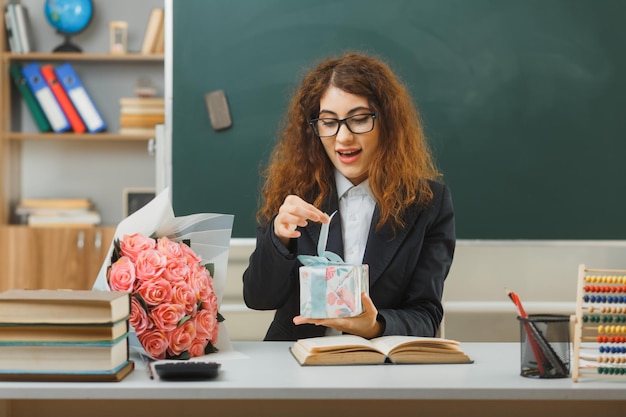 Free photo pleased young female teacher holding and opening gift sitting at desk with school tools in classroom