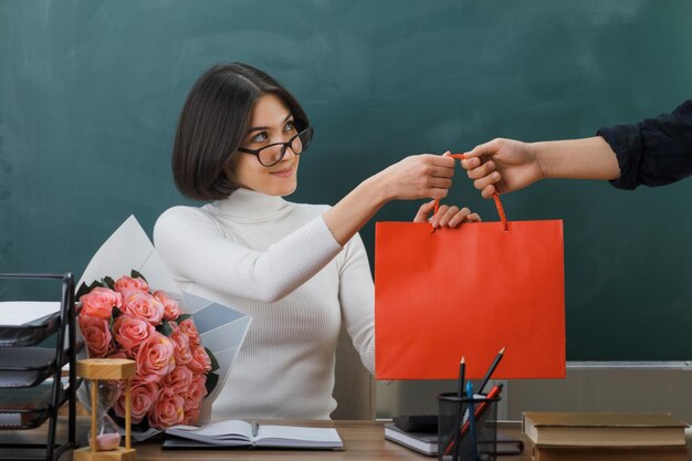 pleased young female teacher received gift bag sitting at desk with school tools in classroom