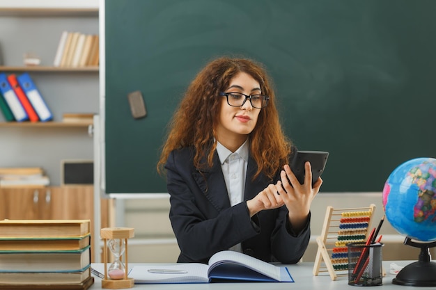 pleased young female teacher wearing glasses holding and looking at calculator sitting at desk with school tools in classroom