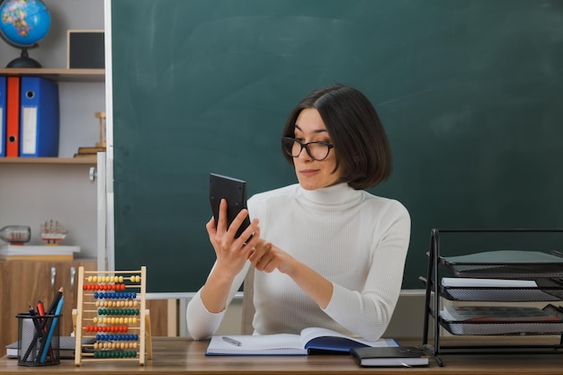 довольная молодая учительница в очках держит и смотрит на калькулятор, сидя за партой со школьными инструментами в классе