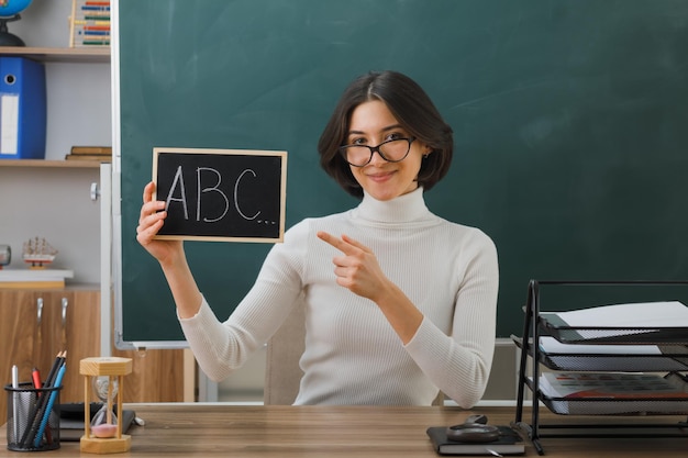 pleased young female teacher wearing glasses holding and points at mini chalkboard sitting at desk with school tools on in classroom