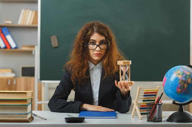 教室で学校の道具と机に座って砂時計を保持している眼鏡をかけている若い女性教師を喜ばせる