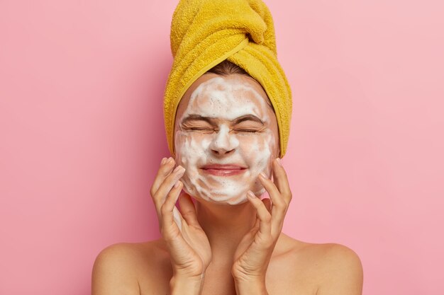 pleased young female touches face skin with soap, keeps eyes closed, washes her face in morning, stands naked against pink wall, has spa treatment. Cleanliness concept