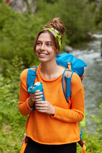 Pleased young female tourist has remarkable hiking trip, enjoys hot drink, holds flask