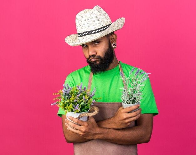Pleased young gardener afro-american guy wearing gardening hat holding and crossing flowers in flowerpot isolated on pink wall