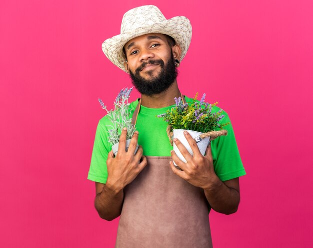 Pleased young gardener afro-american guy wearing gardening hat holding flowers in flowerspot 