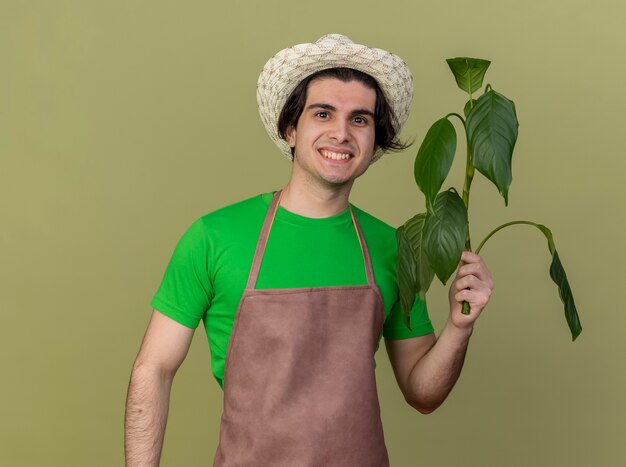 Довольный молодой садовник в фартуке и шляпе держит растение, глядя в камеру с улыбкой на лице, стоя на светлом фоне