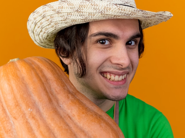 오렌지 배경 위에 서 행복 한 얼굴로 광범위 하 게 웃는 호박을 보여주는 앞치마와 모자에 기쁘게 젊은 정원사 남자