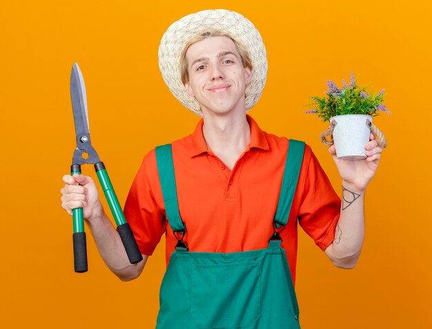 Pleased young gardener man wearing jumpsuit and hat holding hedge clippers