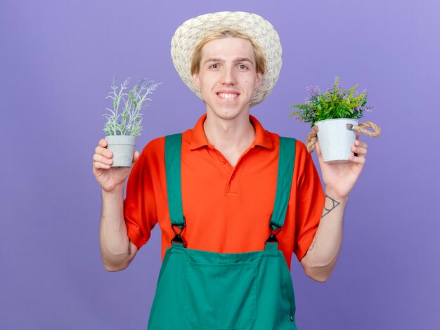 Pleased young gardener man wearing jumpsuit and hat holding potted plants
