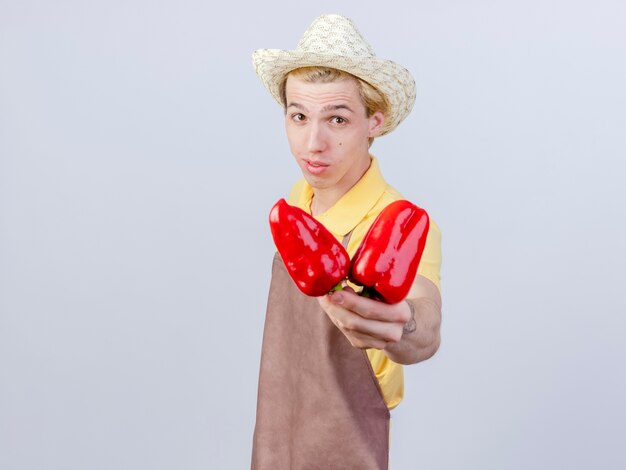 Pleased young gardener man wearing jumpsuit and hat showing red bell peppers with smile on face