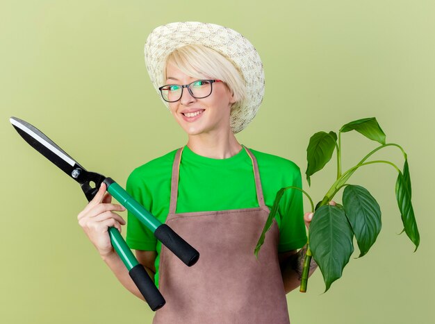 Free photo pleased young gardener woman with short hair in apron and hat holding plant and hedge clippers