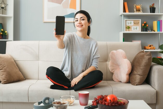 Pleased young girl holding phone, sitting on sofa behind coffee table in living room