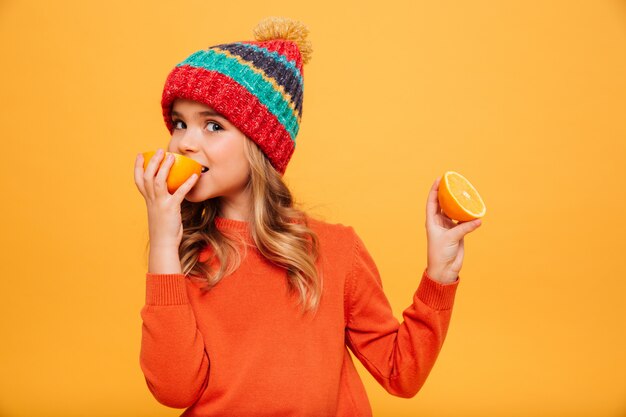 Pleased Young girl in sweater and hat eating orange while looking at the camera over orange