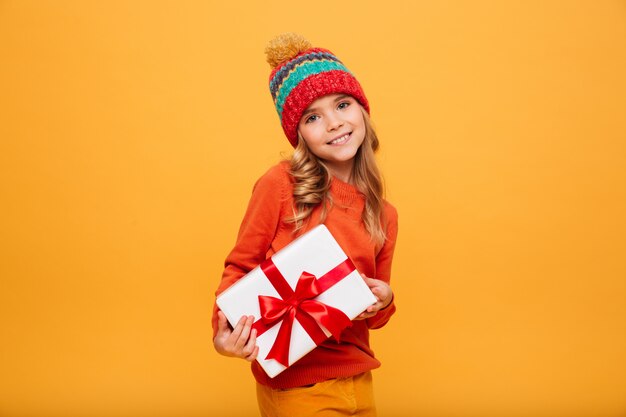 Довольная молодая девушка в свитере и шляпе держит подарочную коробку и смотрит в камеру над апельсином