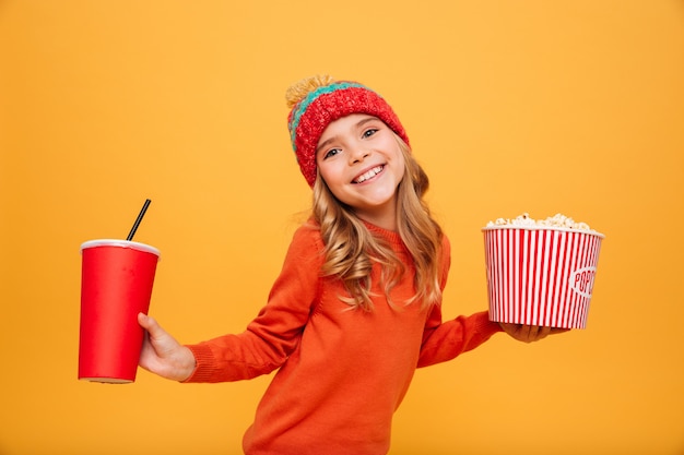 Довольная молодая девушка в свитере и шляпе держит попкорн и пластиковый стаканчик, глядя на камеру над оранжевым