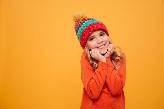 Pleased Young girl in sweater and hat reclines on her arms and looking at the camera over orange