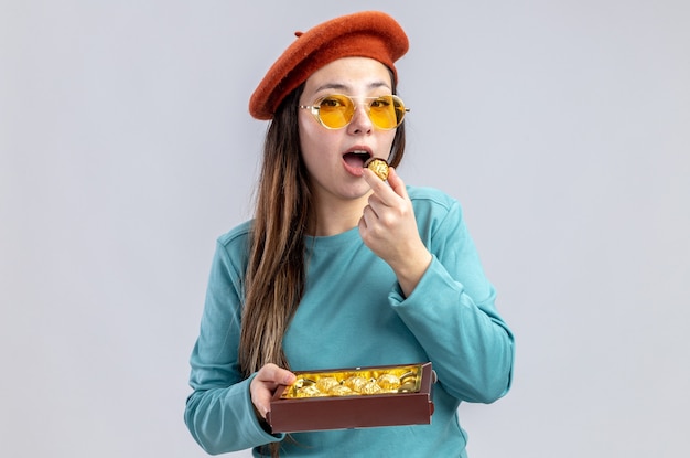 Pleased young girl on valentines day wearing hat with glasses holding box of candies isolated on white background