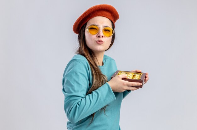 Pleased young girl on valentines day wearing hat with glasses holding box of candies isolated on white background