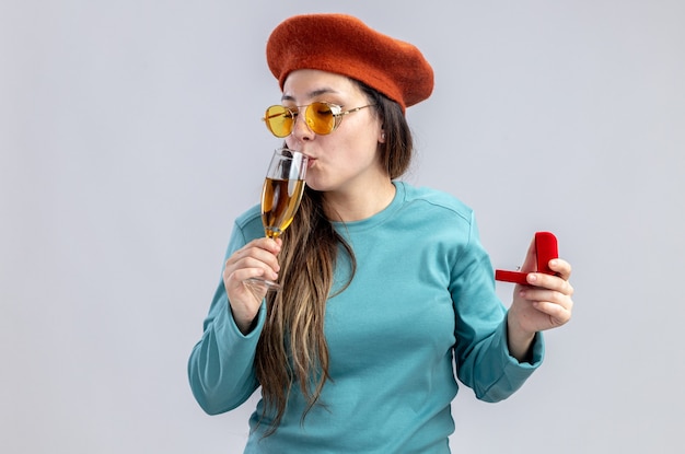 Pleased young girl on valentines day wearing hat with glasses holding glass of champagne with wedding ring isolated on white background