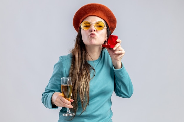 Pleased young girl on valentines day wearing hat with glasses holding glass of champagne with wedding ring showing kiss gesture isolated on white background