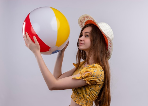 Pleased young girl wearing hat raising beach ball standing in profile view on isolated white space