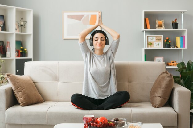 Pleased young girl wearing headphones doing yoga sitting on sofa behind coffee table in living room