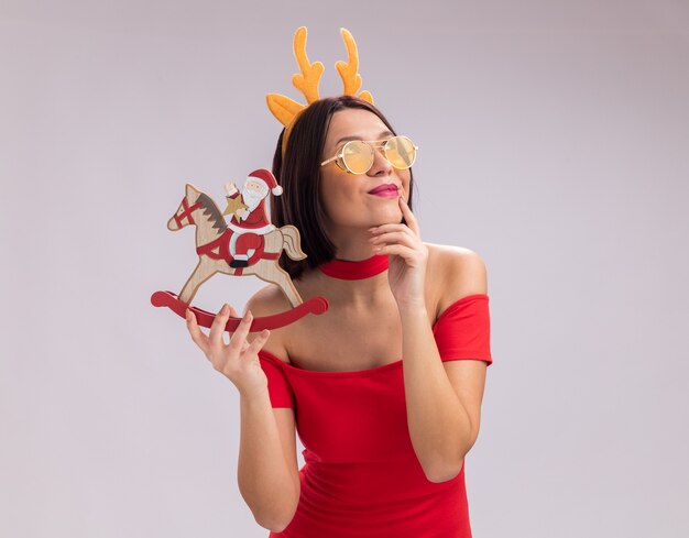 Pleased young girl wearing reindeer antlers headband and glasses holding santa on rocking horse figurine touching chin looking up isolated on white background with copy space