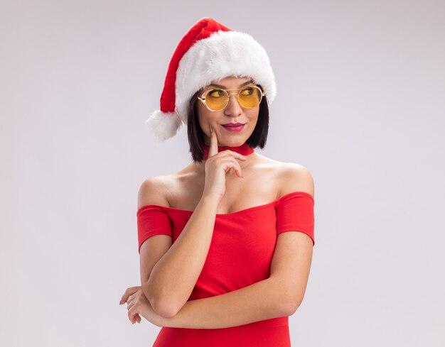 Pleased young girl wearing santa hat and glasses looking at side touching face isolated on white background with copy space