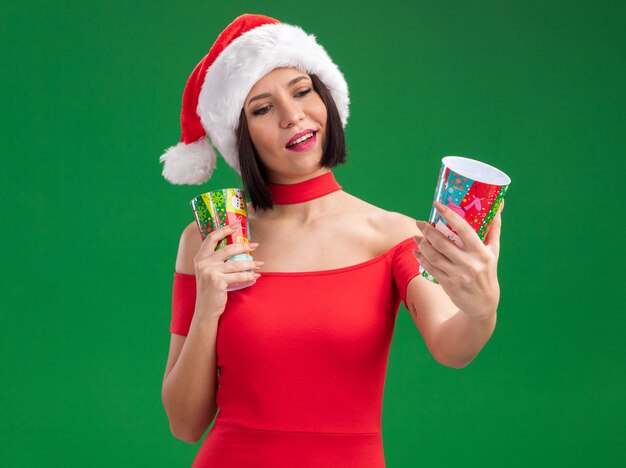 Pleased young girl wearing santa hat holding plastic christmas cups stretching out one looking at it isolated on green background