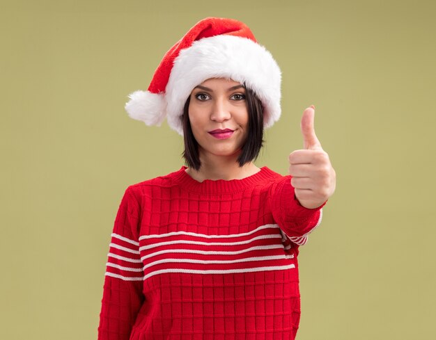 Pleased young girl wearing santa hat looking at camera showing thumb up isolated on olive green background