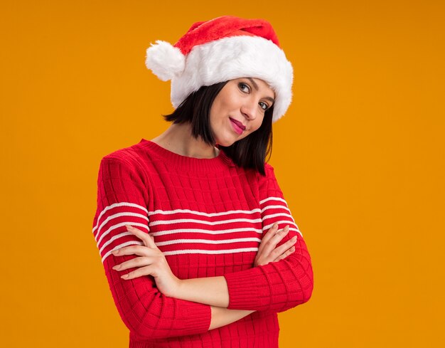 Pleased young girl wearing santa hat standing with closed posture  isolated on orange wall with copy space