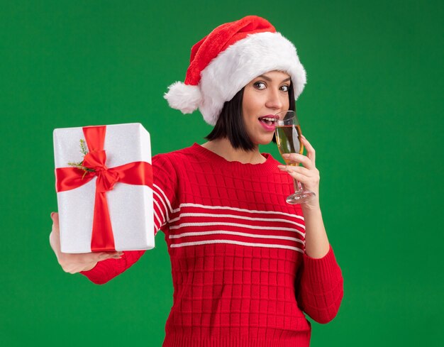 Pleased young girl wearing santa hat stretching out gift package towards camera and drinking glass of champagne looking at camera isolated on green background