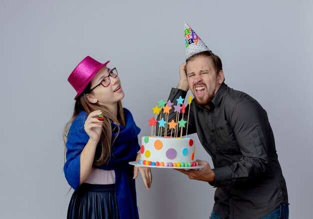 Pleased young girl with glasses wearing pink hat holds whistle looking up and annoyed handsome man in birthday cap holding cake and puts hand on head isolated on white wall