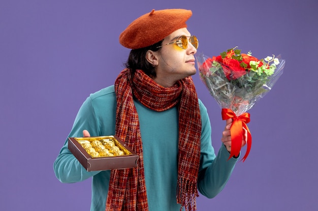 青い背景で隔離の彼の手で花束を嗅ぐキャンディーの箱を保持しているスカーフとメガネの帽子をかぶってバレンタインデーに喜んで若い男