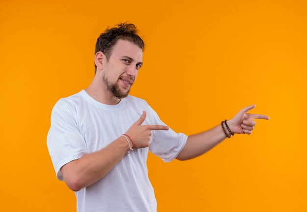 Довольный молодой парень в белой футболке указывает в сторону на изолированном оранжевом фоне