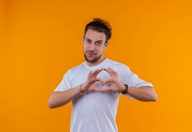 Pleased young guy wearing white t-shirt showing heart gesture on isolated orange background