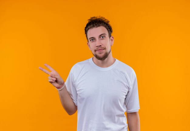 Pleased young guy wearing white t-shirt showing peace gesture on isolated orange background