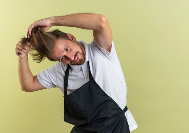 Pleased young handsome barber wearing uniform combing his hair looking at side isolated on olive green wall