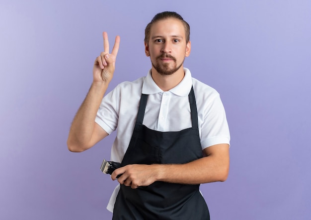 Pleased young handsome barber wearing uniform doing peace sign and holding hair clippers in another hand looking at front isolated on purple wall