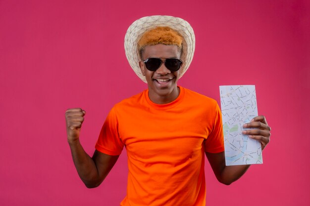 Pleased young handsome boy in summer hat wearing orange t-shirt holding map