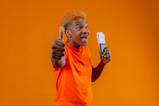 Pleased young handsome boy wearing orange t-shirt holding air ticket smiling cheerfully exited and happy showing thumbs up standing over orange wall