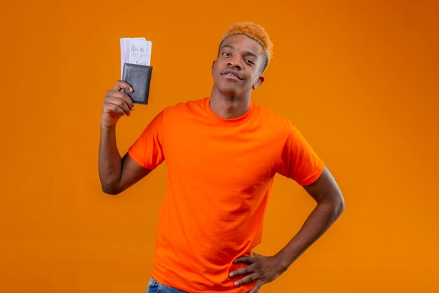 Pleased young handsome boy wearing orange t-shirt holding air ticket smiling confident standing over orange wall