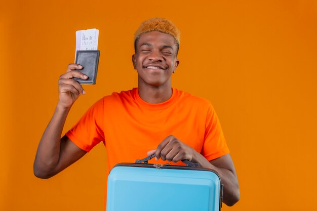 Pleased young handsome boy wearing orange t-shirt holding travel suitcase and airplane tickets smiling happy and exited rejoicing his success standing over orange wall
