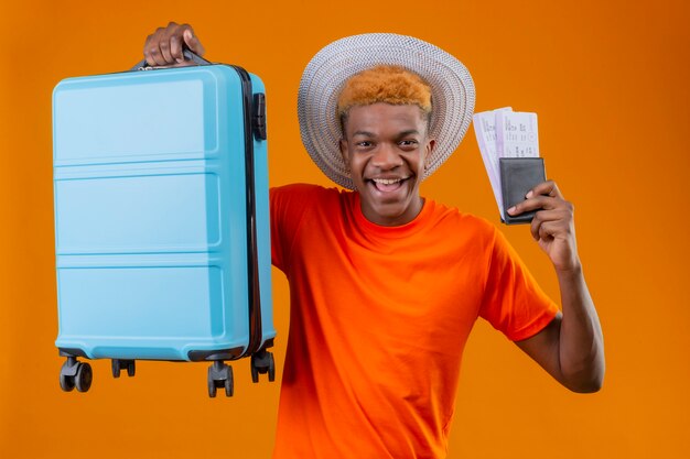 Pleased young handsome boy wearing orange t-shirt holding travel suitcase and airplane tickets