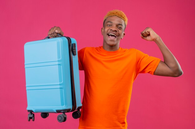 Pleased young handsome boy wearing orange t-shirt holding travel suitcase smiling happy and exited raising fist rejoicing his success standing over pink wall
