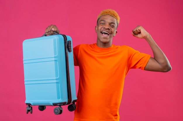 幸せな笑顔の旅行スーツケースを持ってオレンジ色のtシャツを着て満足している若いハンサムな男の子とピンクの壁の上に立って彼の成功を喜んで拳を上げる終了